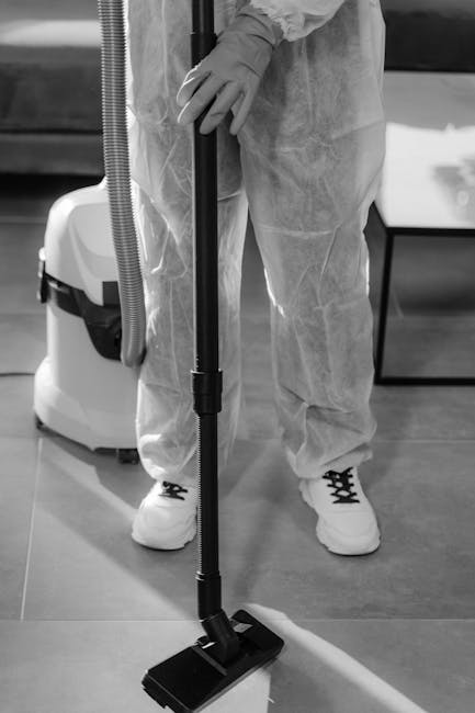 A person dressed in protective clothing, including gloves and a full-body suit, is vacuuming a hard floor surface in a professional cleaning setting. The individual holds a vacuum cleaner with a long 