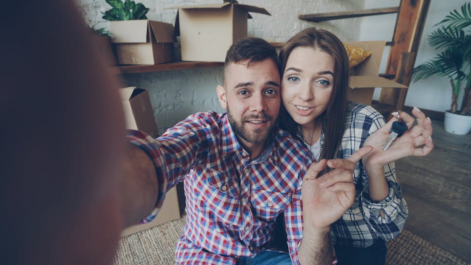 A man and woman sitting on a carpeted floor in a modern, well-lit room, holding up a set of keys. The man is wearing a red and blue checkered shirt, and the woman has long brown hair and is wearing a similar patterned shirt. The background features a wooden shelf with cardboard boxes, a potted plant, and a wall with white bricks, indicating a recent move or a new home. The surface appears clean and tidy, with the focus on the keys and the happy expressions, showcasing the process of moving-in or tenancy handover, relevant to the best end of tenancy cleaning services offered by Cleaner Edgware.
